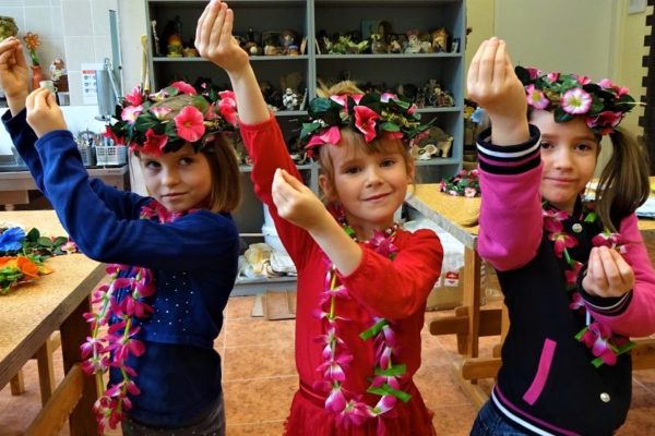 stojące obok siebie trzy dziewczynki w różowych wiankach i z girlandą kwiatów na szyi. Ręce mają uniesione z jednej strony do góry. Połączone palce u dłoni naśladują pąki kwiatowe.