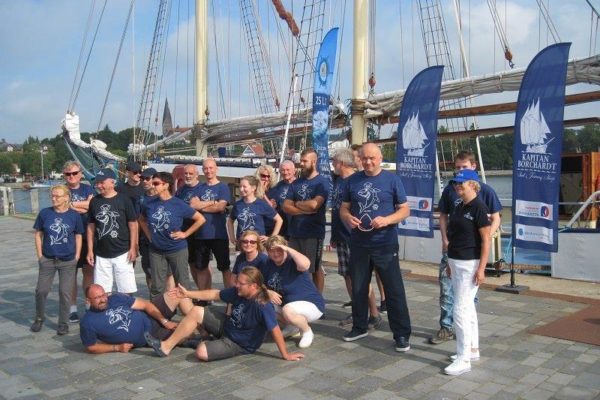Wspólne zdjęcie kadry nauczycieli SKEM wraz z załogą w granatowych koszulkach rejsowych na kei przed żaglowcem