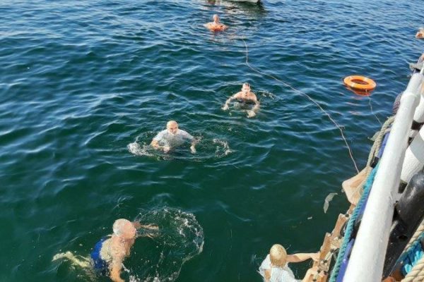 Zdjęcie z burty statku, w morzu kąpie się pięć osób, dwa koła ratunkowe pływają w pobliżu