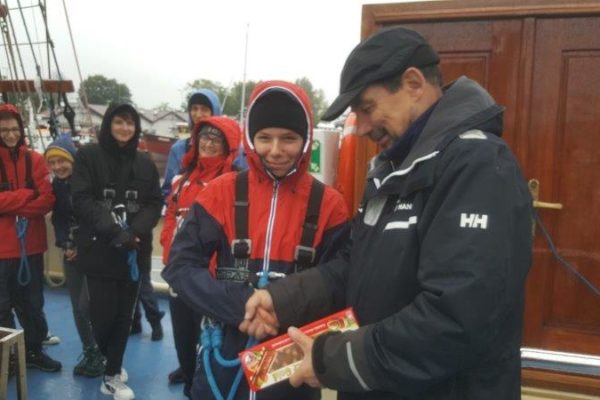 Kapitan wręcza uczestnikowi rejsu tzw. Prezesowi dużą czekoladę z orzechami, w tle 7 uśmiechniętych osób