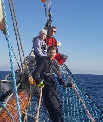 2 uczestników rejsu znajduje się na bukszprycie 1 jest na siatce ochronnej oparty o bukszpryt wszyscy zwróceni twarzami do rufy żaglowca. W tle widać spokojne morze aż po horyzont