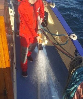 Samuel w czerwonym sztormiaku idzie z wężem strażackim i polewa pokład wodą przy zejściówce dziobowej. Za nim inny uczestnik rejsu przytrzymuje wąż