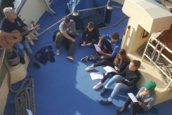 Mechanik Jurek przygrywa na gitarze oparty o burtę a na pokładzie w części dziobowej siedzi 6 uczestników rejsu, którzy trzymają w rękach teksty szant