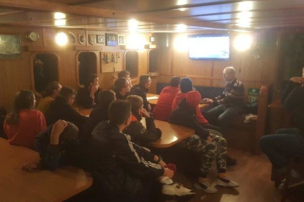 W mesie przy stołach znajdują się uczestnicy rejsu zwróceni w stronę prowadzącego szkolenie kpt Jerzego Szwocha, który stoi przy ekranie telewizora