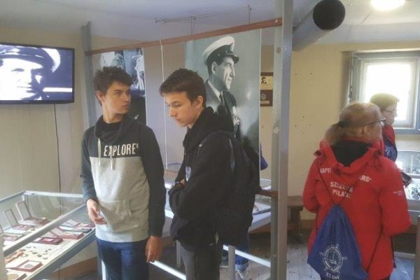 Michał i Antoni oglądają eksponaty oraz zdjęcia w Izbie Pamięci Kpt Karola Borchardta w Muzeum Obrony Wybrzeża w Helu – Wieża Kierowania Ogniem Baterii Szleswig – Holstein w tle Wiesia i Monika opiekunowie