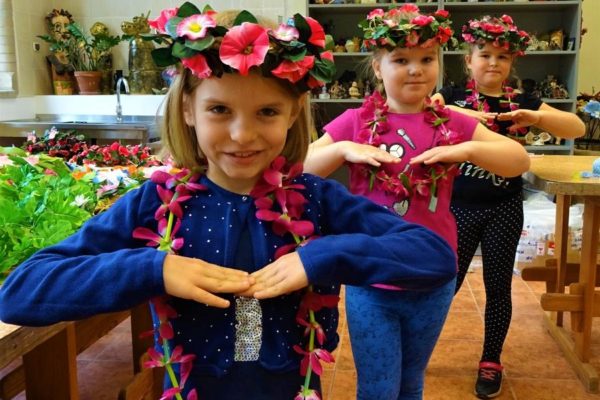 trzy dziewczynki stojące jedna za drugą w pozycji tanecznej. Na głowach mają wianki z różowymi kwiatkami, a na szyi zawieszone girlandy z różowych płatków.