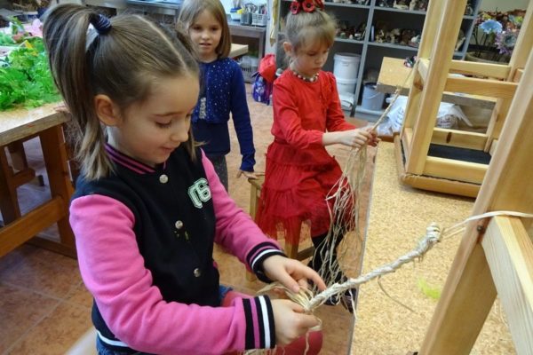 siedzące dziewczęta wyplatają warkocz z rafii. W tle kolejna, trzecia, która obserwuje pracę koleżanek.