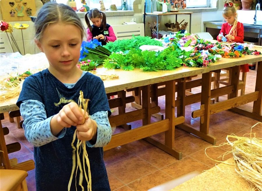 stojąca dziewczynka trzyma w dłoni długie kawałki rafii. W tle dwie dziewczynki wykonują ozdoby z płatków kwiatowych(lei). Na stole rafia, sztuczne kwiaty i liście.