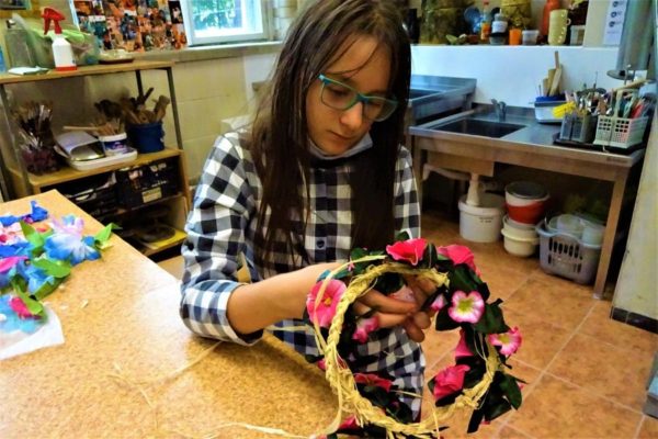dziewczynka trzymająca koło z rafii doczepia do niego różowe kwiaty i liście tworząc wianek. Na stole płatki niebieskich i różowych kwiatów.
