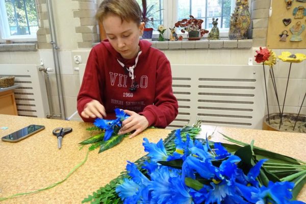 dziewczynka trzymająca koło z rafii doczepia do niego różowe kwiaty i liście tworząc wianek. Na stole płatki niebieskich i różowych kwiatów.