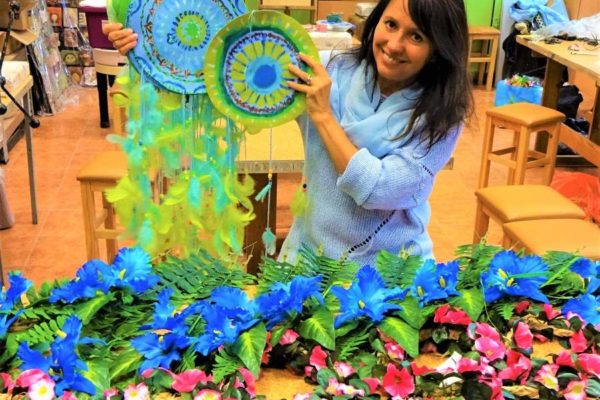 kobieta siedząca przy stole układająca kompozycje z niebiesko-turkusowych kwiatów na zielonych liściach. Na stole leżą bukiety niebieskich kwiatów i liści.