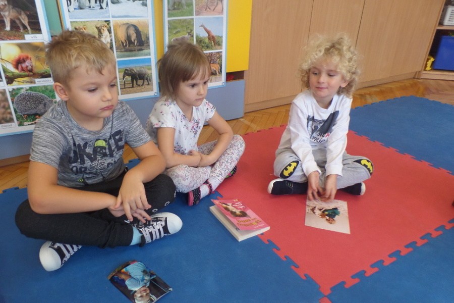 trójka dzieci siedzi na macie, przed dwójką z nich leżą fotografie ich zwierząt, dziewczynka ma przed sobą książkę o zwierzętach