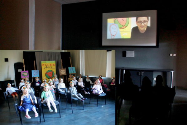 Dzieci siedzą na krzesłach w sali multimedialnej, na ekranie widoczna twarz kobiety, dzieci słuchają bajki