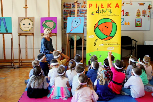 Nauczycielka z grupą dzieci w sali. Dzieci siedzą na macie