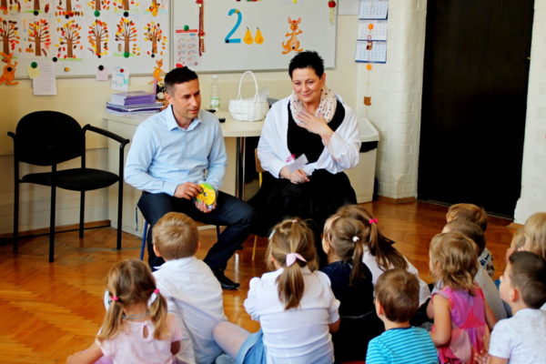 Mężczyzna i kobieta rozmawiają z przedszkolakami. Mężczyzna trzyma w ręku płyty CD z bajką