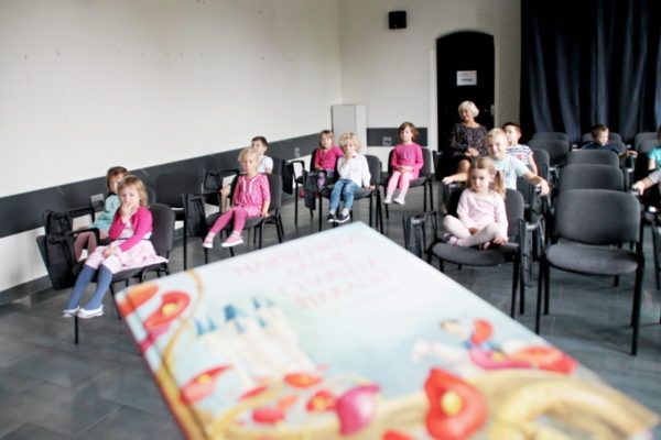 Grupa przedszkolaków siedzi na krzesłach w sali multimedialnej, na pierwszym planie książka