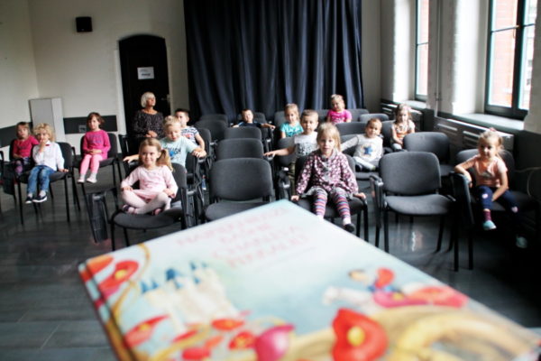 Grupa przedszkolaków siedzi na krzesłach w sali multimedialnej, na pierwszym planie książka