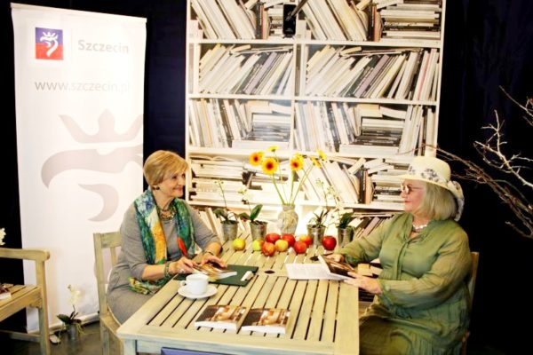 Autorka wraz z prowadzącą spotkanie siedzą przy stole, na stole wazon z kwiatami, jabłka