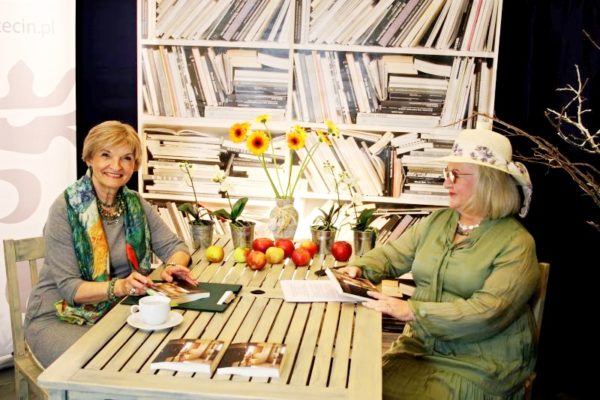 Autorka wraz z prowadzącą spotkanie siedzą przy stole, na nim wazon z kwiatami, jabłka