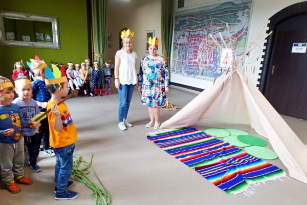 Na pierwszym planie chłopiec rzucający oszczepem, za nim inni przedszkolacy, po prawej tipi i stojące dwie nauczycielki