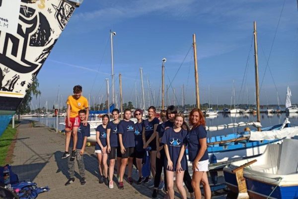 grupa 10-osobowa grupa uczniów Szkolnego Koła Edukacji Morskiej z Technikum Ekonomicznego wraz z opiekunką p. Basią na przystani Centrum Żeglarskiego, w tle zacumowane jachty i maszty