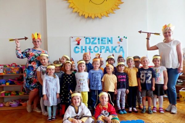 Zdjęcie grupowe - przedszkolaki z dwiema wychowawczyniami, na ścianie napis DZIEŃ CHŁOPAKA