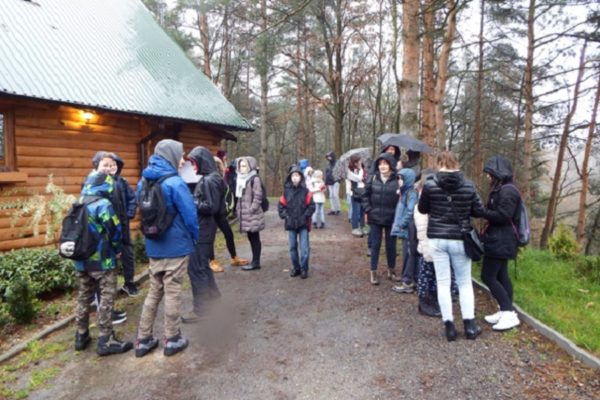 Grupa dzieci z opiekunami stoi obok drewnanego budynku