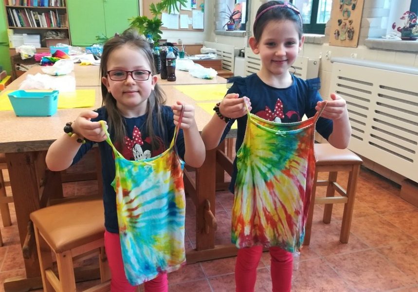 Trzy uśmiechnięte dziewczynki stoją w sali obok siebie trzymając kolorowe, farbowane bluzki