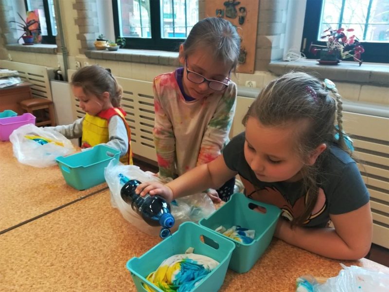 Trzy dziewczynki siedzą przy stole. Przed nimi w pojemnikach zabarwione tkaniny. Jedna trzyma plastikową butelkę z barwnikiem.