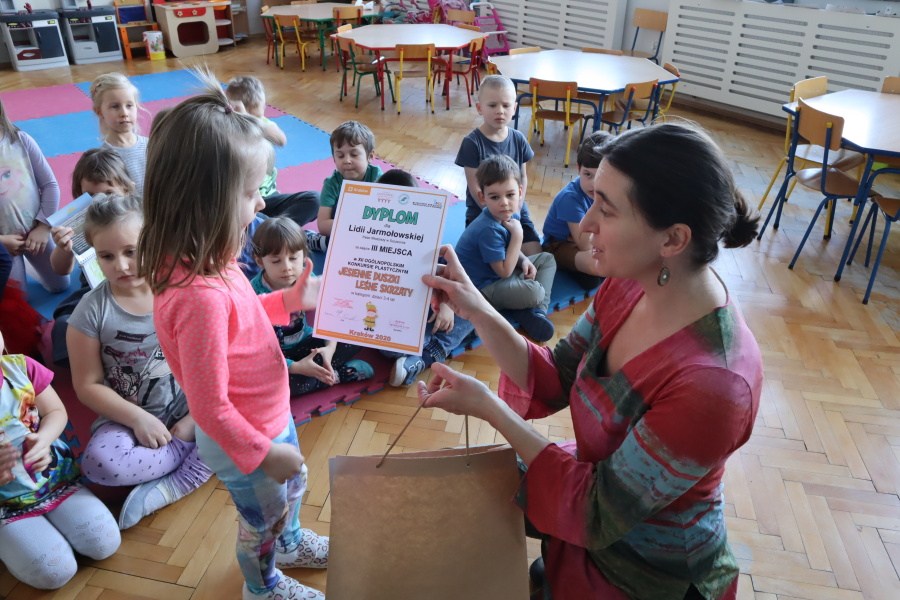 dziewczynka odbiera nagrodę z rąk nauczcielki w tle pozostałe dzieci