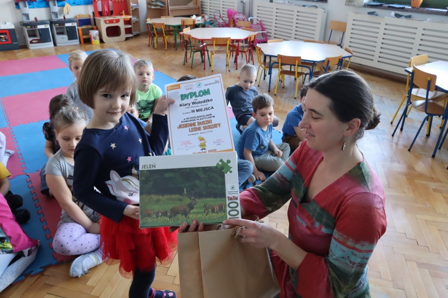 dziewczynka odbiera nagrodę z rąk nauczcielki w tle pozostałe dzieci