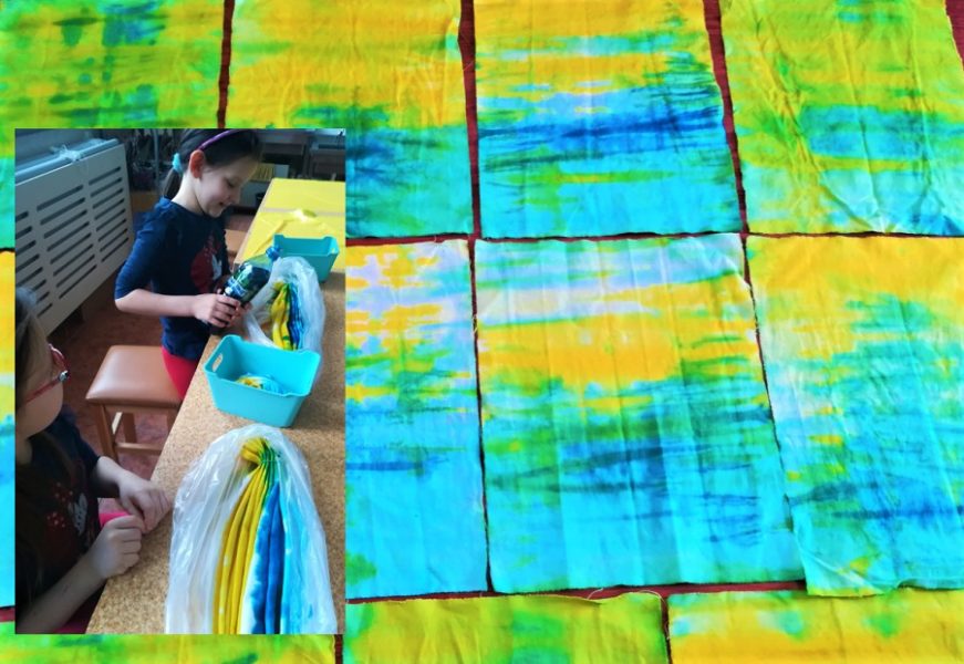 W lewym boku zdjęcia dwie dziewczynki. Jedna siedzi, a druga stoi trzymając butelkę z barwnikiem. Przed nimi w foliowych woreczkach barwna tkanina. Tło stanowią tkaniny pofarbowane na kolor żółto-niebieski