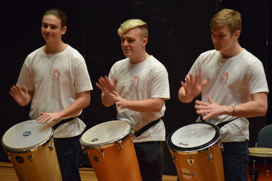 fotografia przedstawia trzech uczniów w białych strojach grających na brazylijskich etnicznych instrumentach.