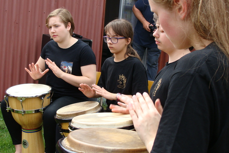 fotografia przedstawia czworo uczniów ubranych na czarno grających w plenerze na afrykańskich bębnach.
