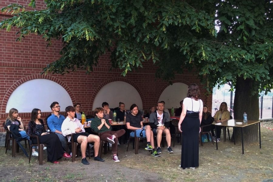 grupa młodych ludzi, przed nimi kobieta stojąca tyłem do fotografa