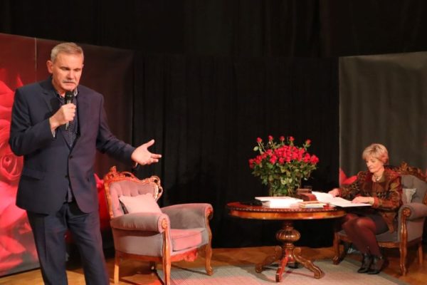 mężczyzna z mikrofonem w dłoni, kobieta siędzi przy stoliku