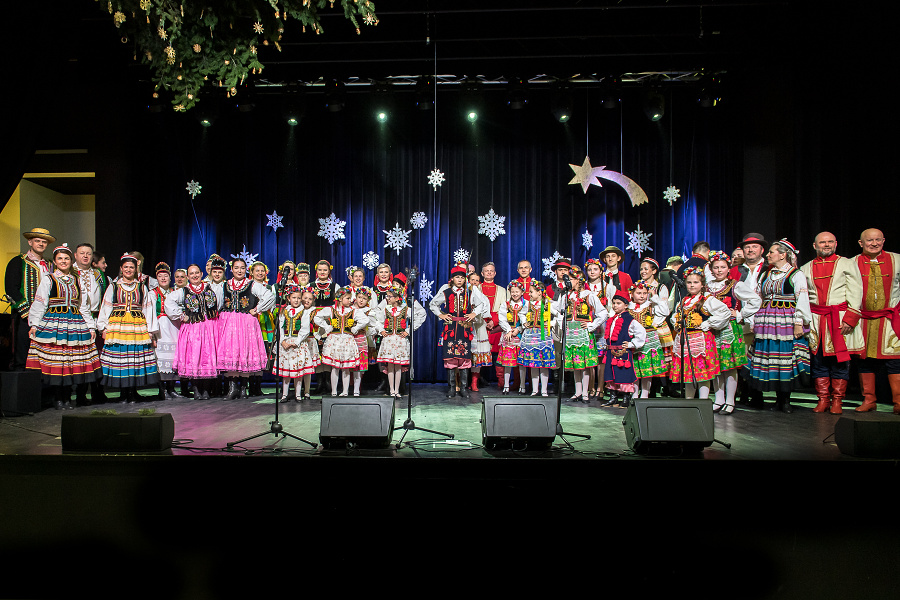  grupę artystów w zróżnicowanych strojach ludowych, stojącą na scenie ze świąteczną dekoracją