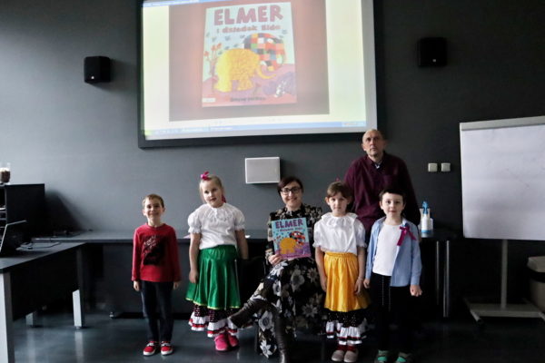 grupa dzieci pozuje do zdjęcia z nauczycielami na tle ekranu na którym jest okładka ksiażki