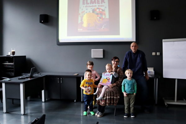 grupa dzieci pozuje do zdjęcia z nauczycielami na tle ekranu na którym jest okładka ksiażki