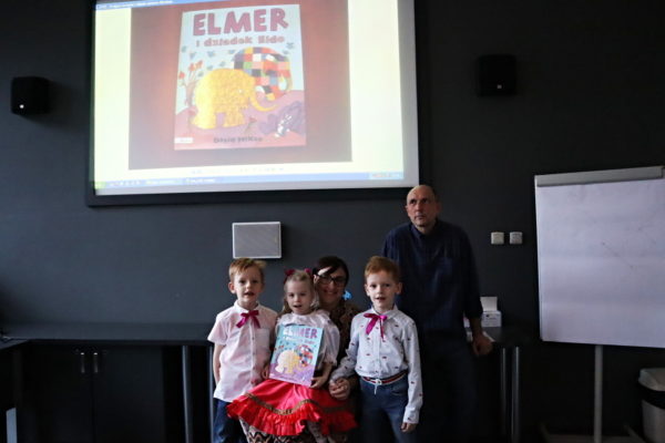 grupa dzieci pozuje do zdjęcia z nauczycielami na tle ekranu na którym jest okładka ksiażki