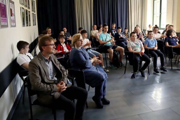 grupa osób zgromaszonych w sali siedzą na krzesłach