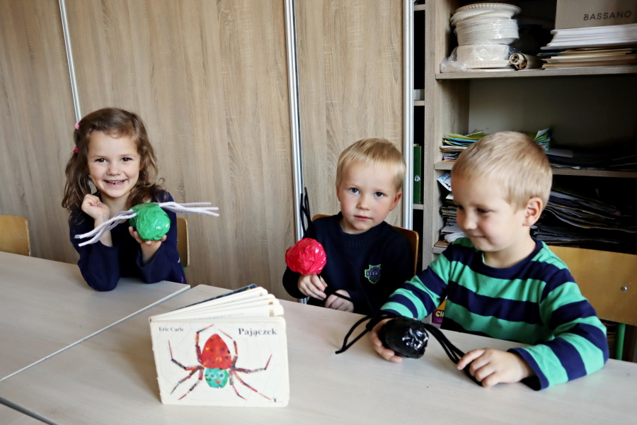trójka dzieci siedzi przy stoliku w ręku trzymają pajączki przed nimi książka