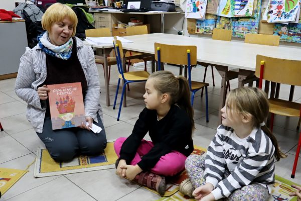 nauczycielka z ksiażką w ręku obok siedzi dwójka dzieci