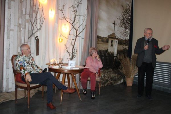 dwie osoby siedzą przy stoliku mężczyna stoi z mikrofonem