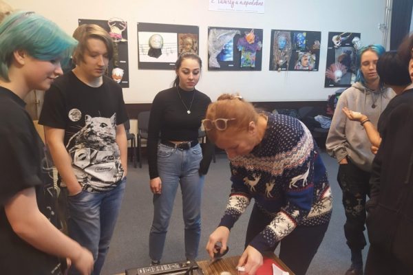 grupa młodzieży stoi wokół nauczycielki pochylonej nad stolikiem