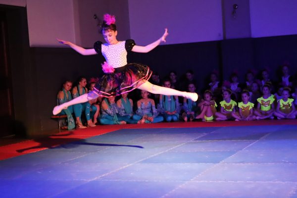 solistka tancerka w podskoku za nią grupa tancerek siedzi