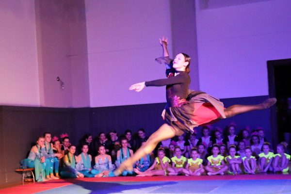 solistka tancerka w podskoku za nią grupa tancerek siedzi