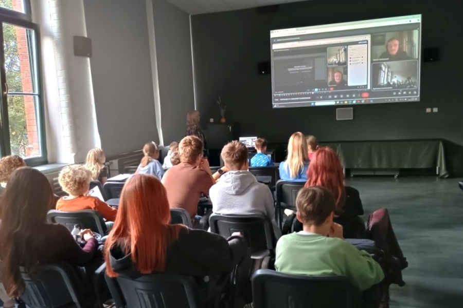 grupa uczestników siedzi na krzesłach w tle ekran na którym spotkanie online