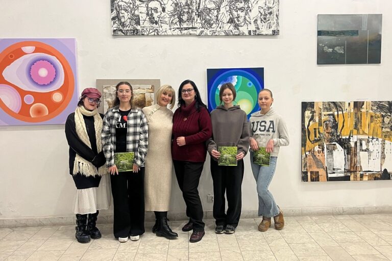 grupa uczestniczek z nauczycielką pozują do zdjęcia na tle obrazów